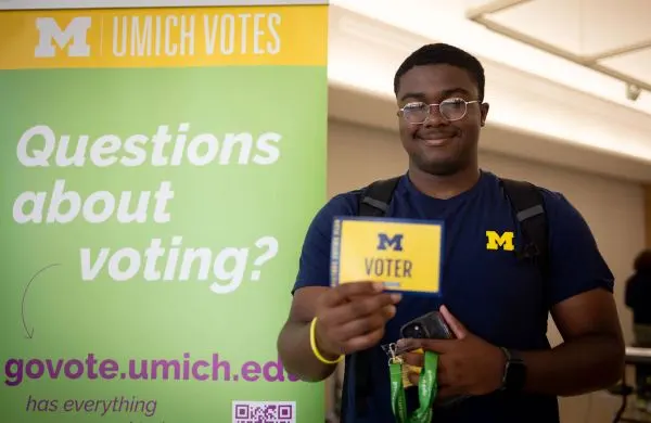 A smiling student walking out of a Umich Votes site carrying a card that says "Voter"