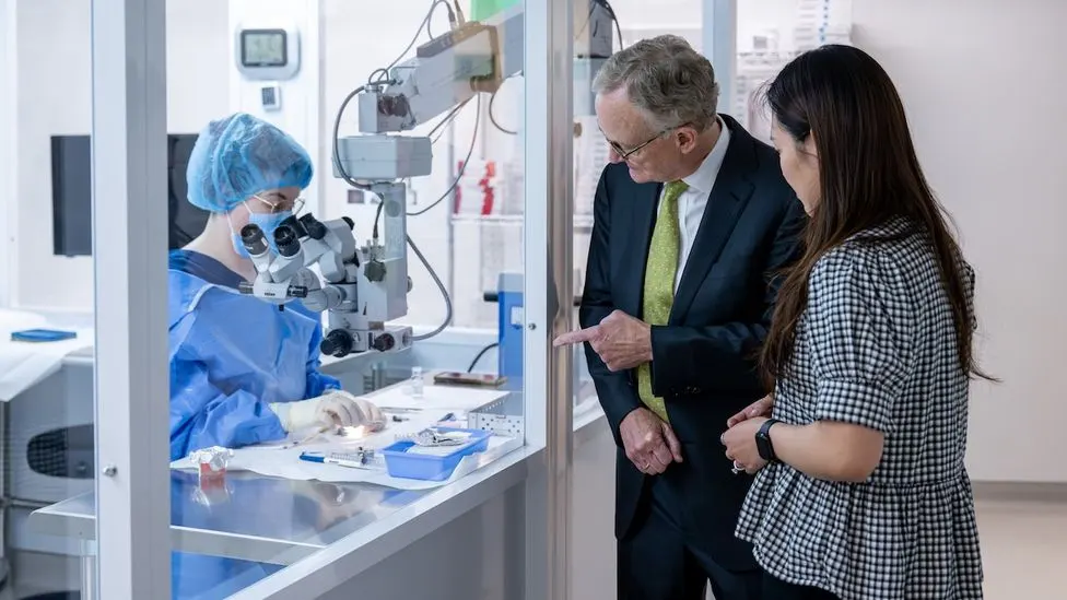 A lab technician in surgical scrubs works at a microscope workstation while two visitors observe and point through a glass partition.A lab technician in surgical scrubs works at a microscope workstation while two visitors observe and point through a glass partition.
