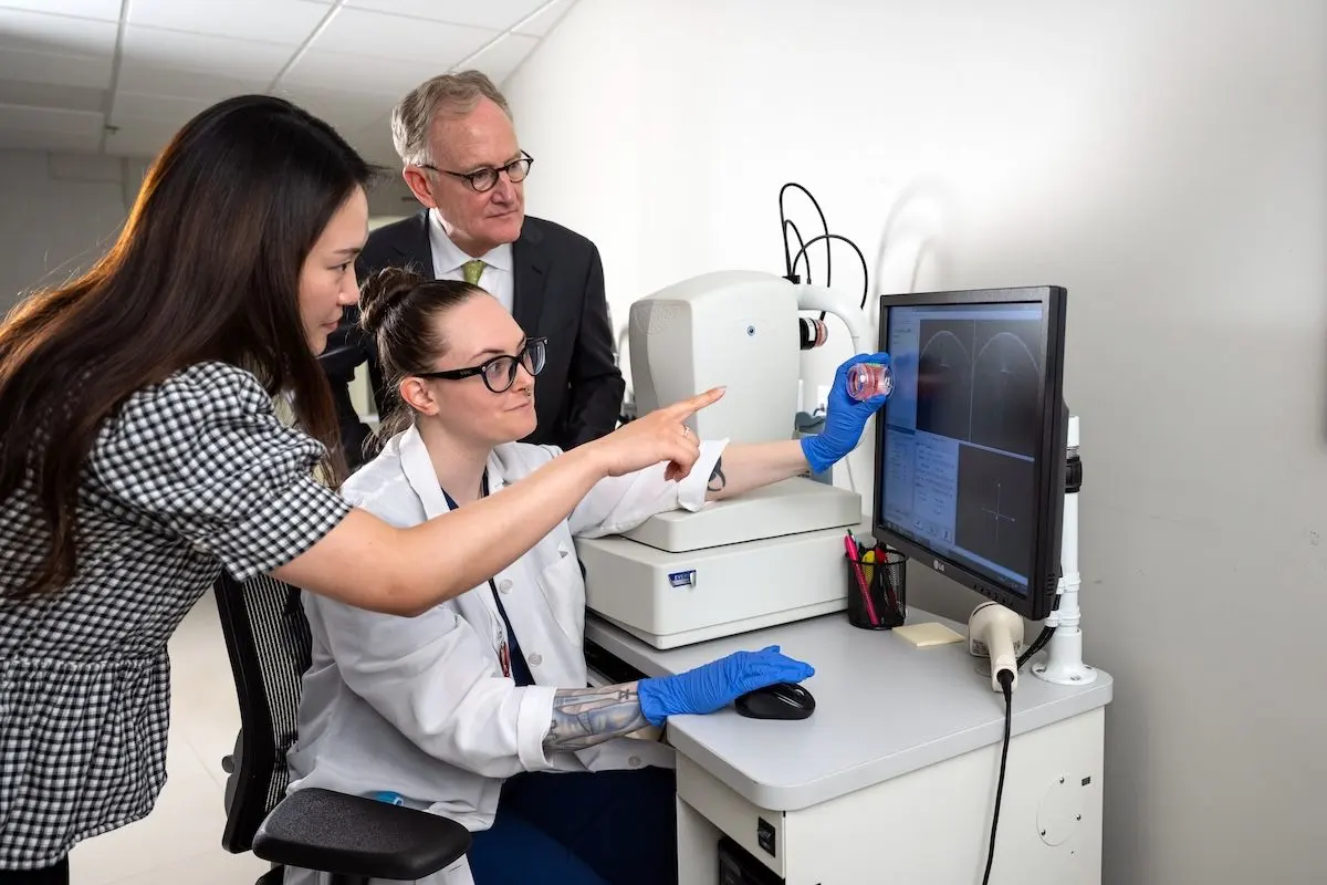 A lab technician in a white coat and blue gloves points to diagnostic imaging on a monitor while two visitors look on beside medical equipment.A lab technician in a white coat and blue gloves points to diagnostic imaging on a monitor while two visitors look on beside medical equipment.