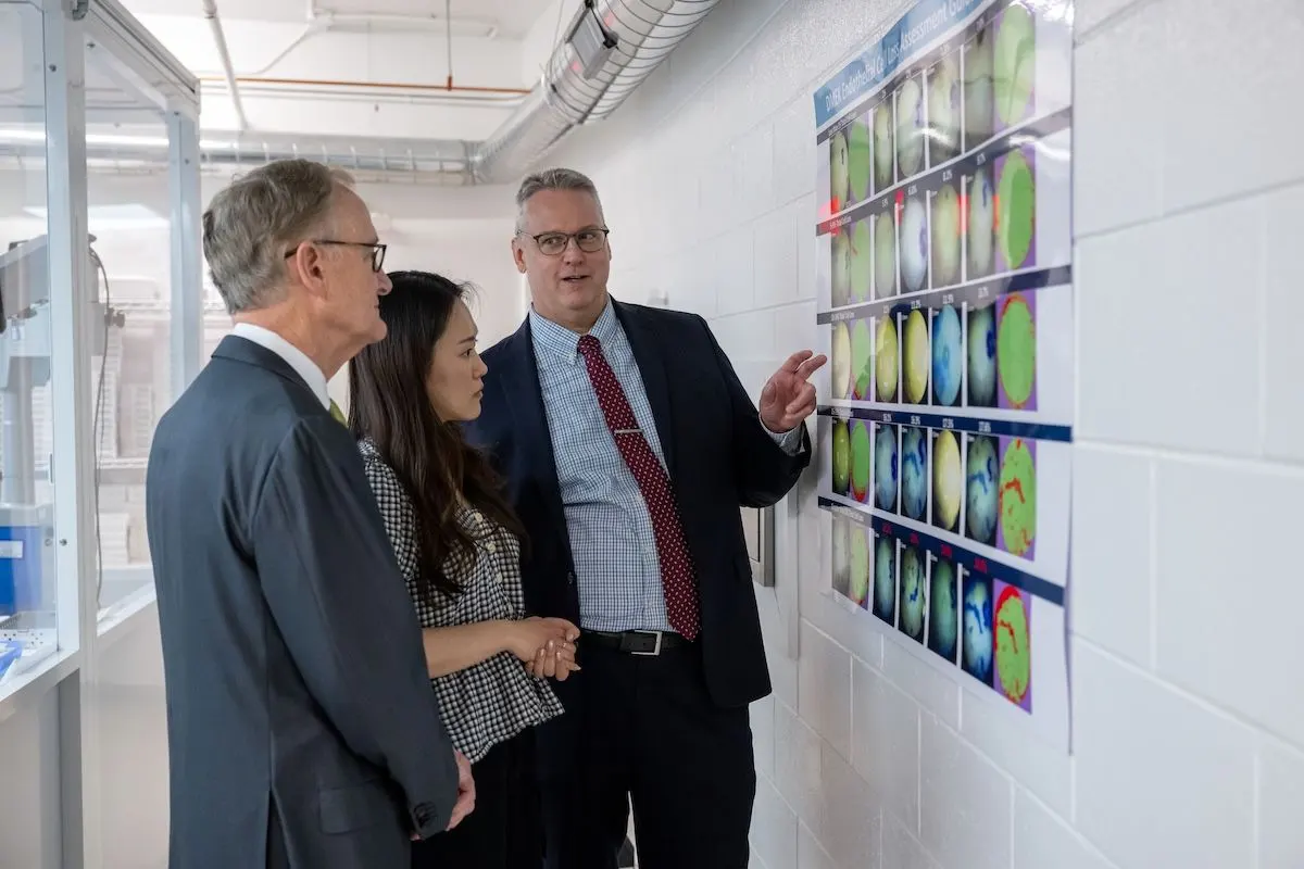 A man in a suit gestures toward a wall display of colorful scientific images while two colleagues look on in a laboratory hallway.